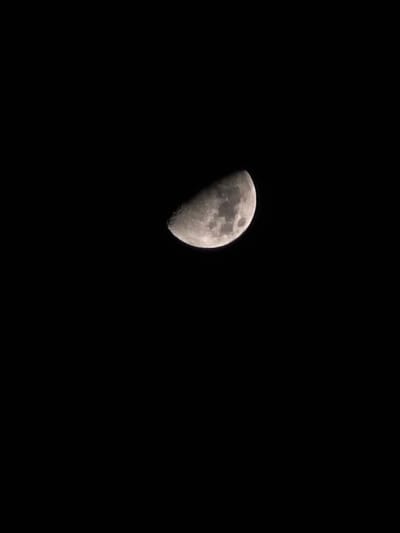 A celestial body identified as the Moon is depicted against a uniform black background, indicative of a night sky, as observed from Trachoni, Cyprus. The Moon is illuminated in a half-phase, presenting as a first or third quarter moon, with its terminator line appearing straight. Surface details including numerous craters and variations in albedo are discernible across the illuminated, grayish-white portion. No other celestial objects, atmospheric conditions, people, or terrestrial features are present in the frame.