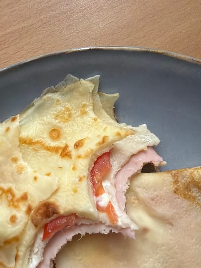 A close-up shot shows a partially eaten savory crepe on a grey plate. The crepe is filled with ham, what appears to be cheese, and diced red tomatoes. The crepe itself is golden brown with some darker, slightly charred spots. The plate is set on a wooden surface. The scene is indoors, likely in a home kitchen. Ożarów Mazowiecki, Poland is not visible in this image.