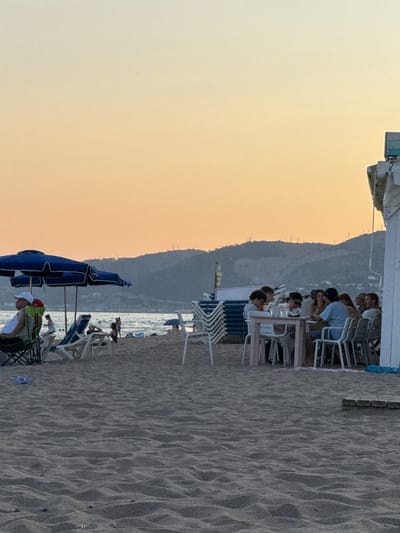 This image depicts a beach scene in Castelldefels, Spain, during what appears to be late afternoon or early evening, given the soft, warm hues of the sky. The setting is a sandy beach with the gentle waves of the Mediterranean Sea visible in the background.

The primary subjects are a group of people gathered at tables set up on the sand, likely at a beachfront restaurant or cafe. They are seated, engaged in conversation, and appear to be enjoying a meal or drinks. The people are diverse, with individuals of various ages and genders. Some are dressed casually, in t-shirts and shorts, fitting for a beach environment.

Scattered around the scene are typical beach objects. There are numerous white lounge chairs, many stacked neatly, suggesting they belong to the establishment or are available for rent. Several beach umbrellas, including a prominent large blue one, provide shade. The sand itself is a textured element, showing the characteristic ripples left by the wind or water. In the distance, a mountainous landscape rises, providing a backdrop to the coastal setting.

The activity taking place is leisurely. People are relaxing, socializing, and dining by the sea. The overall mood seems tranquil and pleasant, indicative of a typical vacation or leisure day at the beach. The time of day is suggested by the warm, soft light illuminating the sky, casting a gentle glow over the scene. There are no visible text elements in the image.