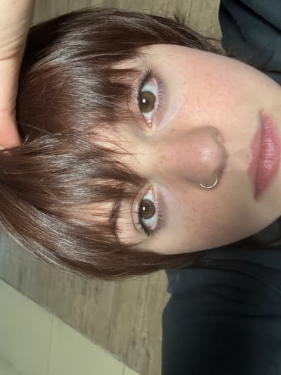 An upside-down selfie shows a person with brown eyes and brown hair. The hair is styled with bangs. The individual has a nose piercing and pink lips. Their skin is fair and freckled. They appear to be wearing eye makeup. The background is blurry, with a hint of what looks like a beige wall and a wooden floor.