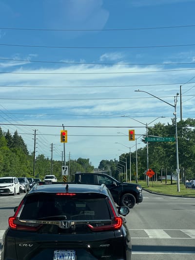 This vertical, wide-angle, eye-level shot captures a traffic scene in Richmond Hill, Canada, on a clear, bright day, likely mid-morning or early afternoon. The foreground is dominated by the rear of a black Honda CR-V, with its brake lights illuminated. A line of other vehicles, including white cars and a black SUV, are visible behind it, suggesting a traffic jam or a wait at an intersection. The road is paved, with white pedestrian crosswalk markings clearly visible.

Above the cars, a prominent green street sign reads "ELGIN MILLS RD WEST." Traffic lights are also visible, with at least one showing a red signal, reinforcing the idea of a traffic delay. Overhead power lines crisscross the sky, a common sight in this area. Further back, lush green trees line the sides of the road, indicating a suburban or semi-rural setting.

In the background, various signs are present. One orange diamond-shaped sign warns of "CONSTRUCTION 2 km," and another displays a speed limit of "38." A billboard with a portrait and text is partially visible in the distance. The presence of "Uber" on the back of the CR-V suggests ride-sharing services are active in the area. The overall impression is a typical daytime commute or travel experience in a Canadian suburb, marked by traffic and roadwork.