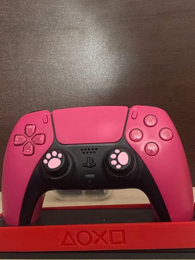 A bright pink PlayStation 5 controller with black accents is displayed on a red controller stand. The controller features a D-pad on the left and face buttons on the right, with paw-print themed analog sticks. The stand has embossed PlayStation button symbols on its front. The background appears to be a textured, brown surface. The image is shot from a slightly elevated angle, focusing on the controller and its stand.