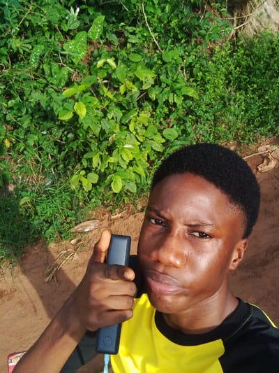 The image shows a young man of African descent holding a black power bank with a blue charging cable attached. He is wearing a yellow and black t-shirt and is standing outdoors. The background consists of dense green foliage and reddish-brown soil, suggesting a natural or semi-natural environment. The location could be in or around Orlu, Nigeria, based on the prompt, but without additional evidence, this cannot be definitively confirmed from the image itself.