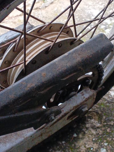 The image displays a close-up, low-angle view of the rear wheel and drive components of a motorized two-wheeled vehicle, situated on a mottled concrete or paved surface in Kyauk Hsoke, Myanmar. The central element is a light-colored metal wheel hub, possibly aluminum, featuring concentric ridges and secured by numerous dark, rust-colored spokes. A dark, tubular metal chain guard, heavily corroded with rust and encrusted with dirt and debris, extends diagonally across the mid-ground, partially obscuring a rusty metal chain and a toothed sprocket positioned beneath it. A metallic swingarm component, showing significant rust and secured by a visible hexagonal bolt, extends into the lower right portion of the frame. All visible metallic components exhibit advanced stages of corrosion, pitting, and accumulated surface grime. No individuals, actions, or interactions are depicted. The background is a blurred, light-gray and brown textured surface, consistent with worn pavement or concrete.