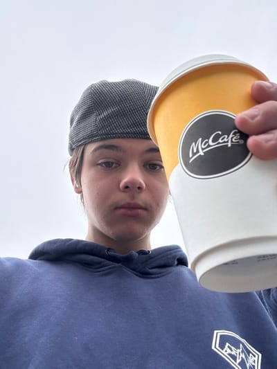 An individual, identified as a young male, is depicted in a close-up, low-angle shot, presenting a McCafé branded cup. He wears a grey herringbone flat cap and a navy blue hooded sweatshirt with a white geometric mountain graphic on the lower right chest. Small silver stud earrings are visible in both earlobes. His expression is neutral as he looks directly into the camera. His right hand holds the yellow-orange and white McCafé cup, which is positioned prominently in the foreground, obscuring part of his face. The background consists solely of an overcast, light grey sky. No specific environmental markers are present to identify a precise location within the city of Kirklees, United Kingdom.