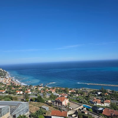 The image shows a panoramic view of the coastal town of Ospedaletti, Italy, with the Mediterranean Sea in the background. The sky is a clear, vibrant blue, with a faint, dotted line of what appears to be contrails from an aircraft stretching horizontally across the upper portion of the frame.
The foreground features a greenhouse structure with a glass roof and metal frame, situated on a dry, grassy hillside. Beyond the greenhouse, the landscape descends towards the town, revealing a mosaic of terracotta-tiled roofs on houses and buildings scattered amongst lush green trees and vegetation. The town itself hugs the coastline, with a crescent-shaped beach and a series of wave-breaking structures extending into the turquoise and deep blue waters of the sea. A long, man-made breakwater, constructed of large rocks, extends out into the sea.
Further inland, a swimming pool with bright blue water is visible, surrounded by paved patios and a low wall. Various residential buildings with the characteristic red-tiled roofs of the region are nestled into the hillside, some appearing quite substantial with balconies and terraces. A road runs parallel to the coastline, with some vehicles visible. The overall impression is a sunny day in a picturesque Italian coastal town, with evidence of both residential life and agricultural activity (the greenhouse). There are no people visible in the image.