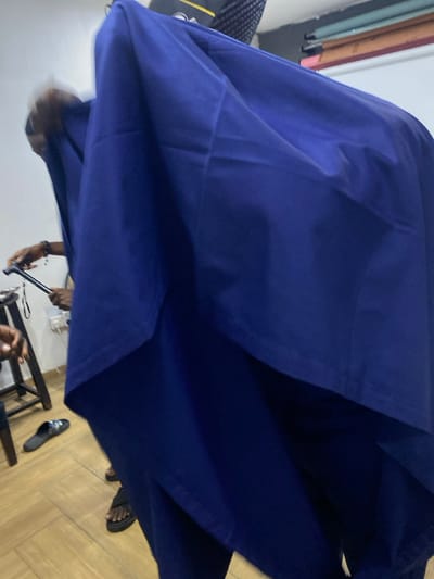 A dark blue garment dominates the frame. The clothing item obscures much of the background, leaving glimpses of a room. One can discern a wooden floor, a rack with multiple fabric rolls, a partial view of a bag, and the legs of people nearby. One man is using a black cane, while another wears sandals. A black sandal is visible on the floor.