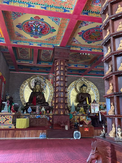 The image depicts the interior of a Buddhist temple in Việt Trì, Vietnam. The scene is dominated by two large, seated Buddha statues, one on the left and one on the right, each adorned with golden halos. In front of the statues, the floor is covered with a red carpet, and there is a wooden altar area with various offerings and decorations. To the right, a tall, wooden shelf structure is filled with numerous smaller Buddha figurines. The ceiling is elaborately decorated with colorful, intricate patterns and motifs. The overall atmosphere suggests a sacred and ornate religious space.