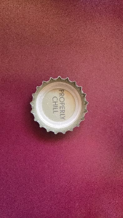 A silver bottle cap lies on a textured, deep red surface. The cap's interior is light gray and stamped with the words "PROPERLY CHILL" in a simple, dark gray font. The cap's edges are crimped, standard for bottle caps. The red background exhibits a subtle gradient, darkening towards the top-right and lightening towards the bottom-left. The lighting in the scene is even, providing moderate contrast.