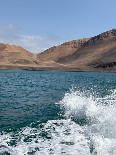 The image shows a choppy turquoise ocean in the foreground with white foam and spray from a boat's wake. In the background, arid, brown hills rise steeply from the water's edge under a clear blue sky with some wispy clouds. A small lighthouse or tower structure is visible on the crest of one of the hills. This is likely the area around Isla San Lorenzo in Callao, Peru, known for its rugged, dry landscape and coastal waters.