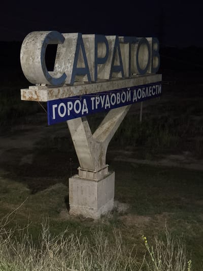 The image shows a weathered concrete sign at night, likely marking the entrance to a city or town. The sign features large, three-dimensional letters that spell out "САРАТОВ" (Saratov) in a light gray color with blue outlines. Below this, a blue rectangular banner with white text reads "ГОРОД ТРУДОВОЙ ДОБЛЕСТИ" (City of Labor Valor). The sign is supported by a robust concrete structure that appears aged, with visible signs of wear and tear, including chipping paint and rust stains. The surrounding environment is dark and appears to be a grassy area with dry, sparse vegetation in the foreground. The overall lighting suggests artificial illumination from an unseen source, casting shadows on the ground. There are no people visible in the image.