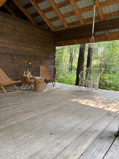 The image depicts a wooden porch in Bol'shaya Rechka, Russia, likely part of a rustic cabin or dacha. The porch is furnished with two wooden lounge chairs, a small foldable table with a hookah and bottles on it, and a tree trunk serving as a stool. A macrame hammock chair is suspended from the porch roof, overlooking a dense forest. The wooden floorboards are weathered, and sunlight casts dappled shadows. The environment is natural and serene, suggesting a peaceful retreat.