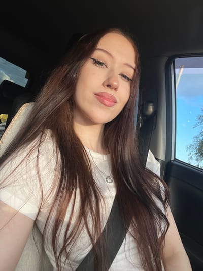 The image is a selfie taken from a low angle inside a car. The main subject is a young woman with long, dark brown hair, which is spread across her shoulders and chest. She is wearing a white t-shirt with a small design on the left side of her chest. Her face is partially visible, with her eyes closed and her lips slightly parted, suggesting a relaxed or possibly sleepy expression. She has winged eyeliner and pink lipstick. A black seatbelt is visible across her chest.

The setting appears to be daytime, with sunlight filtering into the car from the open sunroof and the side window. The sky visible through the sunroof is blue with some clouds, and trees can be seen in the distance, indicating an outdoor location, likely a rural or suburban area given the context of Gavrilova, Russia. The interior of the car is visible, including dark upholstery and the roof lining.

The overall mood of the image is casual and personal, typical of a selfie. The lighting suggests it is either late afternoon or early morning, given the warm, directional sunlight. There is no visible text in the image.
