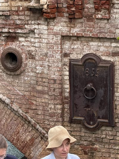The image shows a weathered brick wall with a metal plaque featuring the year "1855" and the letters "TN". A ring hangs from the plaque. To the left, a circular opening is visible in the brickwork. In the foreground, two people are partially visible. One person wears a light-colored hat and a white shirt, and appears to be speaking. The other person, on the far left, is wearing glasses. The overall scene suggests an outdoor environment, possibly a historical site or an older structure in Verkhoturye, Russia.