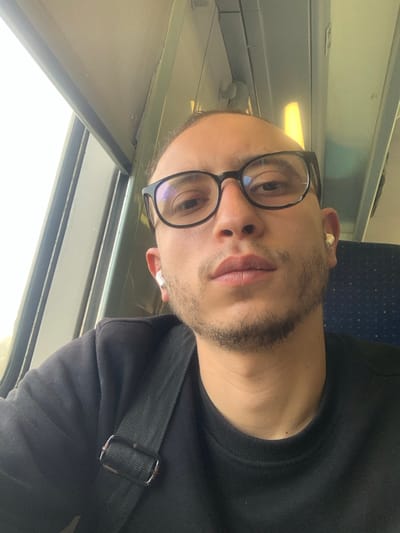 A male individual is depicted in a close-up, head-and-shoulders portrait taken inside a vehicle, likely a train, within the city of Sidi Slimane, Morocco. The subject is light-skinned, has short or pulled-back dark hair, wears black-framed rectangular eyeglasses, and has a short beard or stubble. He is wearing a black long-sleeved shirt or sweatshirt, with a black textile shoulder strap of a bag visible across his left shoulder. White wireless earbuds are present in both ears; the left earbud appears to be an Apple AirPod, while the right has a similar but slightly less defined appearance. The individual gazes directly forward, maintaining a neutral expression. To his left, a large window reveals a bright, undifferentiated sky outside, suggesting daytime. The vehicle's interior features light-colored wall panels and overhead, a textured ceiling panel with a warm yellow strip light. A dark blue, patterned seatback is partially visible to the right of the subject.