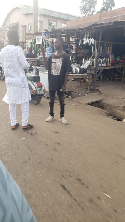 The image depicts a street scene in Kawo, Nigeria. The foreground shows two men standing on a paved surface. One man is wearing a white patterned garment while the other is in a dark t-shirt and dark trousers. A motorcycle is parked between them. Behind the men is a shop selling various metal items. A POS sign is also visible near the shop. A building with windows is present in the background.