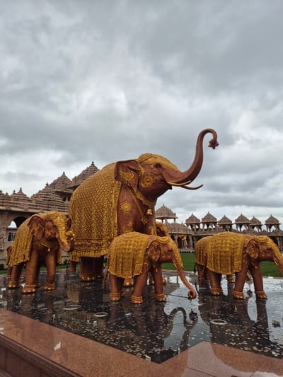 In Robbinsville Township, United States of America, a collection of elephant statues, adorned with ornate golden coverings, stand on a reflective surface filled with scattered coins. In the background, a large, intricately designed temple structure is visible under a cloudy sky. The statues appear to be made of a reddish-brown material and are decorated with detailed patterns. The reflective surface suggests it is a water feature or a highly polished floor. The overall scene conveys a sense of grandeur and cultural significance associated with the temple complex.