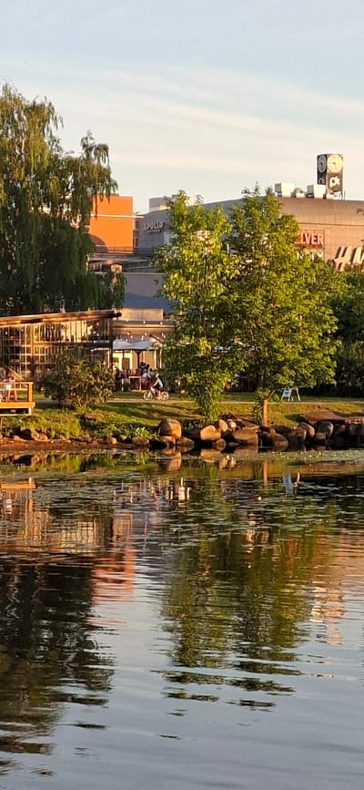 In Pärnu linn, Estonia, the image captures a waterside scene with a modern building complex in the background, illuminated by the warm glow of late afternoon sun. The building features a prominent sign displaying "APOLLO" and another with the word "IVER," suggesting a commercial or entertainment venue. Above these, a large clock tower with Roman numerals is visible. In the foreground, a tranquil body of water reflects the sky and the surrounding greenery. The water's surface is dotted with lily pads, and a small bird is perched on a rock along the water's edge. A paved walkway runs along the water, with a seated individual visible on a wooden deck to the left. Further along the walkway, a group of people are gathered under umbrellas outside a glass-enclosed structure, possibly a restaurant. A person is seen riding a bicycle on the walkway. Lush green trees and foliage line the bank, creating a natural buffer between the water and the urban structures. The overall atmosphere is serene and inviting, blending natural elements with contemporary architecture.