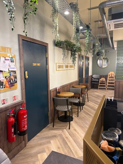 Here is a detailed description of the image:
**General Description:**
The image depicts an interior space with a mix of what appears to be a staff area and a customer area. The view is angled to the right, giving a perspective that elongates the space.
**Location Details:**
There's not enough information in the image to determine if the location is in Mole Valley, United Kingdom.
**Subjects and Objects:**
* **Doors:** There are two dark blue doors, one with the inscription "STAFF ONLY." These doors seem to lead to restricted areas.
* **Seating Area:** Several modern chairs with wooden or gray seating are visible. These chairs are likely part of a café or waiting area.
* **Decorations:**
* Greenery: Foliage and decorative plants are hanging from above, adding a natural aesthetic.
* Mirror: A round mirror is placed on one of the walls, likely for decorative purposes.
* Sign: A sign says "COUPLE 1000-PEOPLE"
* **Furniture:** A counter or shelf area is seen in the foreground.
* **Fire Safety Equipment:** Two red fire extinguishers are placed near the "STAFF ONLY" door.
* **Notice Board:** A notice board with various papers and documents is affixed to the wall beneath the "STAFF ONLY" door.
**Actions and Interactions:**
There are no visible actions or interactions taking place. The scene appears to be static.
**Environment:**
* **Flooring:** The floor has a wood-like pattern, possibly laminate or wood paneling.
* **Lighting:** Overhead lighting fixtures are present, and natural light seems to be entering the space.
* **Overall Ambiance:** The ambiance seems to be a mix of modern and rustic, with the greenery adding a touch of nature to the interior.
**Overall Impression:**
The image portrays a functional yet aesthetically pleasing interior space, likely a business with staff and customer areas. The presence of safety equipment and signage suggests a professional and organized environment.