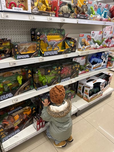 The image shows a child at a store shelf stocked with dinosaur toys, likely in Korobovo, Russia. The child is wearing a green jacket with a fur-lined hood and a brown patterned hat. They are bending down to look at the toys on the lower shelves. The shelves are filled with various dinosaur toys in boxes, mostly from the Jurassic World and Robo Alive brands. The prices of the toys are visible on the shelf tags, ranging from 1,099 to 3,999. The floor is tiled with light-colored square tiles.