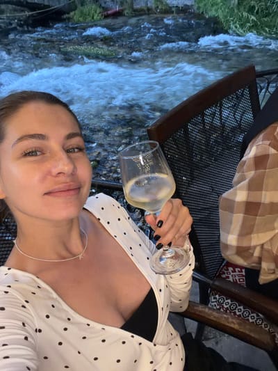 The media file shows a woman taking a selfie in an outdoor setting. She is holding a wine glass filled with a white beverage and wearing a white polka-dot top with a black undershirt. Her nails are painted black. The background features a stream of water flowing over rocks, with some greenery to the left. To the right, there's a glimpse of a table and chairs, suggesting a dining or seating area, possibly at a restaurant or cafe. A checkered blanket is visible on one of the chairs. The lighting suggests it might be evening or a shaded area. The overall mood appears relaxed and leisurely. The woman's facial expression is soft and pleasant. No visible text is present in the image. The location context provided is Travnik, Bosnia and Herzegovina.