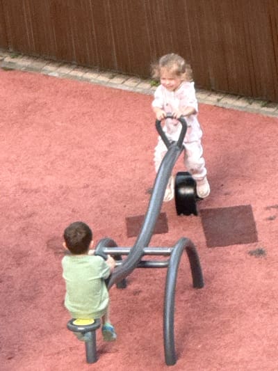 Two young children are playing on a playground in what appears to be a residential courtyard. The ground is covered in a reddish-pink, textured surface, possibly rubberized safety surfacing common in playgrounds. To the left, a wooden fence or wall runs diagonally across the frame.

In the foreground, a child wearing a light green shirt and blue shoes is lying on their back, straddling a metal see-saw. Their head is on the left side of the see-saw, and their legs are extended towards the right. The see-saw itself is a curved metal structure supported by vertical poles.

Further back, another child, a girl, is sitting on the opposite end of the see-saw, wearing a white long-sleeved shirt and pink pants. Her hands are resting on a black object attached to the see-saw. Her hair is blonde and appears to be short.

The scene is shot from a high-angle perspective, looking down at the children. There are a few dark square-shaped shadows on the ground, suggesting the presence of overhead structures or trees. The lighting appears to be natural daylight, and the overall mood seems to be one of playful activity. No visible text is present in the image. The location context of Сухарево, Russia, indicates an urban residential setting.