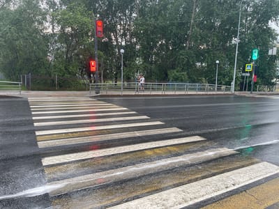 This media file depicts a pedestrian crossing on a wet asphalt road. The crossing has alternating white and yellow stripes. In the background, there is a metal fence and lush green trees. Traffic lights are visible, with one showing a red "walk" signal for pedestrians, and another showing a green light for vehicles. A pedestrian crossing sign is also present. The road appears wet, suggesting recent rain or damp weather. The sky is overcast, indicating a cloudy day. The scene conveys a sense of an ordinary urban environment, likely during the daytime given the lighting conditions. The image was taken in Khanty-Mansiysk, Russia. There is no discernible text in the image.