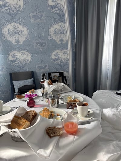The image displays an indoor hotel room, contextually located in Deauville, France, depicting a breakfast setting post-consumption. A round table draped in a white tablecloth occupies the foreground and midground, covered with items. Visible foodstuffs include a silver basket with assorted bread slices, one partially eaten, a plate with pastry remnants and butter pats, a single pancake, and three small jars of preserves. Beverages consist of a partially consumed glass of pink liquid (likely juice) and a white cup with a dark residue on a saucer, accompanied by a silver teapot. A small magenta vase containing a single pink orchid is centrally placed on the table. To the left, a blue upholstered armchair with a cushion holds a dark object. The wall behind the table is covered in light blue wallpaper featuring white toile de Jouy patterns depicting classical scenes. In the background, a Nespresso-style coffee machine and an electric kettle are visible on a surface. To the right, a window with bright natural light is partially obscured by dark grey curtains, and a white, rumpled bed indicates recent occupancy. No individuals are present in the scene.