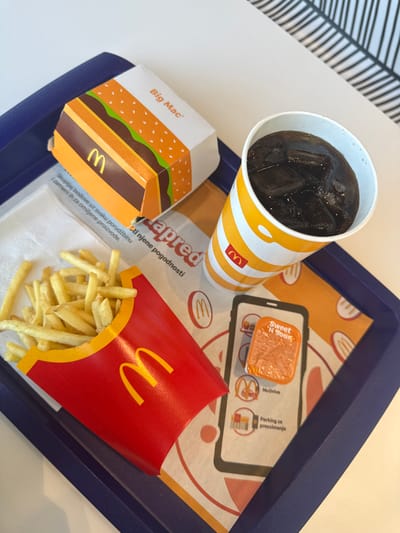 The image shows a McDonald's meal on a blue plastic tray. The tray contains a Big Mac in its box, a drink with ice, a serving of French fries in a red carton, a sachet of Sweet 'N Sour sauce, and promotional materials. The food is placed on a white napkin. The table is white, and there is a black and white striped pattern visible in the upper left corner. The presence of Serbian text (e.g., "apred", "skupljaj bodove", "Parking za preuzimanje") suggests the meal is in Serbia. Given the available information, it can be determined that the meal is likely in a McDonald's restaurant within the Ub Municipality, Serbia.