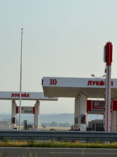 An outdoor, rotated shot depicts a Lukoil gas station situated alongside a multi-lane road, under a clear, bright sky. The station features several pump islands and canopies, predominantly white with red accents. The brand name "ЛУКОЙЛ" (Lukoil) is prominently displayed in red Cyrillic letters on tall signs and the side of a pump structure.
To the right, a gray metal guardrail separates the gas station area from the asphalt road, which has a white line marking its edge. Patches of green grass and some dry vegetation grow along the roadside and behind the guardrail. In the background, beyond the station, hazy hills or mountains are visible under the pale sky.
One pump island clearly displays "Diesel" in large letters, indicating fuel type. Another red sign on a pump island shows "100" and "ecto" with additional Cyrillic text below, which appears to be "ЗА ВСЕ ПОВЕЧ", likely part of an advertising slogan. Small advertising posters with images and text are visible on the pump structures. A person is partially visible near the pumps. A tall, slender white pole with a small red tip stands in the foreground, separate from the main station structure. The scene is illuminated by daylight, suggesting a clear day in Vodenichane, Bulgaria.