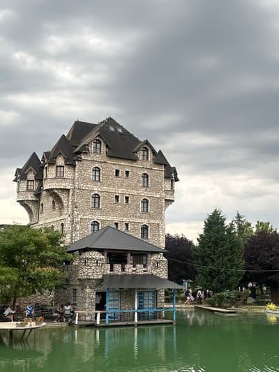 This image depicts a stone building resembling a castle situated next to a tranquil, green body of water. The building features multiple levels with dark pitched roofs, numerous windows, and decorative stone accents. In the foreground, the water reflects the overcast sky and the surrounding greenery. Several people are visible in the scene, some seated at tables near the water's edge, while others walk along a wooden pathway. Trees and bushes add to the naturalistic environment. The overall atmosphere is serene and picturesque. This appears to be the "Etno selo 'Stanišići'" in Bijeljina, Bosnia and Herzegovina.