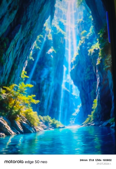 The image depicts a serene natural scene featuring a tranquil body of water flowing through a canyon or gorge. Sunlight streams down from above, illuminating shafts of light that pierce the shadowy depths of the rocky cliffs. Lush green foliage and trees cling to the sides of the canyon, adding vibrant color to the scene. The water is a clear, brilliant blue, reflecting the light and the surrounding environment. On the lower right, text indicates the image was captured with a Motorola Edge 50 Neo, a 24mm lens at f/1.8, a shutter speed of 1/50s, ISO 802, on July 29, 2026. There are no people or any specific identifiable landmarks suggesting the location is Zlataritsa, Bulgaria.