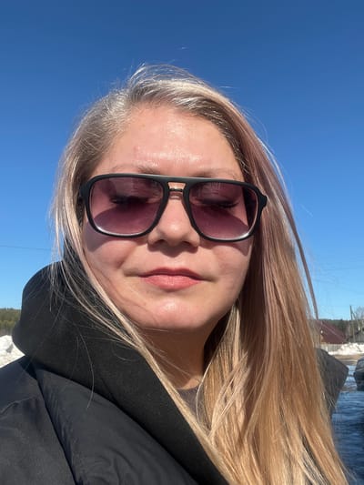 A close-up portrait captured outdoors in Сундур, Russia, features an adult female subject facing forward. She has long, straight blonde hair with noticeable gray roots, worn down around her shoulders. She wears black-framed sunglasses with dark, possibly purple-tinted lenses, and a black hooded jacket or top. Her lips are closed, displaying a light pink hue. The bright blue sky dominates the upper background, with faint utility lines visible extending across the middle-right. Below the horizon, patches of snow are present on the ground, alongside a dark tree line on the left and residential structures with reddish-brown roofs on the right. A dark-colored object, possibly a vehicle, is partially visible on the far right, and the ground in the lower right foreground appears wet or reflective, potentially from melted snow. A faint white contrail is discernible in the upper right quadrant of the sky.
