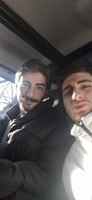 The image depicts two males inside a vehicle, located within the city of Grenoble, France. Both individuals are looking towards the camera, suggesting a self-portrait.

The male on the left, foreground, has dark hair, a full beard, and a mustache. He wears a dark, possibly black, puffer-style jacket, with a seatbelt visible across his chest. Strong sunlight illuminates his left side.

The male on the right is partially visible, cropped on the right side of the frame. He has dark hair and stubble, wearing a light-colored, possibly white or light grey, hooded jacket. His lips are slightly pursed.

The interior of the vehicle shows a grey headliner and dark structural elements overhead, possibly part of a sunroof frame. Through a window on the left side, bare tree branches are visible under bright daylight, indicating an outdoor, possibly winter or early spring, environment.
