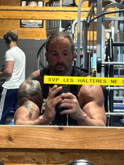 The image captures a mirror selfie taken by a muscular man inside what appears to be a gym or a home workout area in Jemelle, Belgium. The man, with a beard and short dark hair, is positioned centrally, looking directly into the camera of his smartphone, which he holds up. His face shows signs of exertion, with sweat visible on his forehead and around his eyes. He is shirtless, revealing a well-defined, muscular physique and extensive dark tribal-style tattoos on his left arm, shoulder, and side of his torso. His right hand is holding the phone, and his left hand is visible, also tattooed.
The man is reflected in a large mirror, which dominates the scene. A prominent yellow vertical strip of tape is affixed to the mirror, covering part of the reflection. On this tape, black uppercase French text is visible, reading "SVP LES HALTERES NE S". This phrase likely continues to form a complete instruction, such as "SVP LES HALTERES NE PAS LAISSER PAR TERRE" (Please do not leave dumbbells on the ground), indicating a gym rule.
Behind the man, also reflected in the mirror, is the interior of the gym. Various pieces of exercise equipment, including metal racks and weights, are visible in the background, suggesting an active workout environment. To the lower left of the frame (as if rotated upright), a second person is partially visible. This individual is wearing a white t-shirt, dark shorts with white stripes, and over-ear headphones, appearing to be engaged in their own workout. The mirror itself is framed by a light brown wooden border on the right side of the image. The lighting appears to be artificial, common in indoor gym settings, and the overall atmosphere suggests a focused and active workout session.