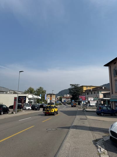 The image captures a street scene in Balerna, Switzerland, on a clear day with a bright blue sky. The perspective is from street level, looking down a road lined with commercial buildings and parked cars. On the left side of the road, there's a row of shops, including one with a sign that reads "CARROZZERIA" and "ASIA THAI TAKE AWAY." Several parked cars are visible in front of these establishments. A prominent yellow SUV with a bicycle rack carrying two bicycles is driving down the road in the right lane, moving away from the camera. Further down the road, a dark blue car is parked on the right side. Various buildings, including a multi-story structure with balconies, are visible on both sides of the street. Trees and rolling hills form the background. The overall impression is of a typical suburban or town street with ongoing daily activity.