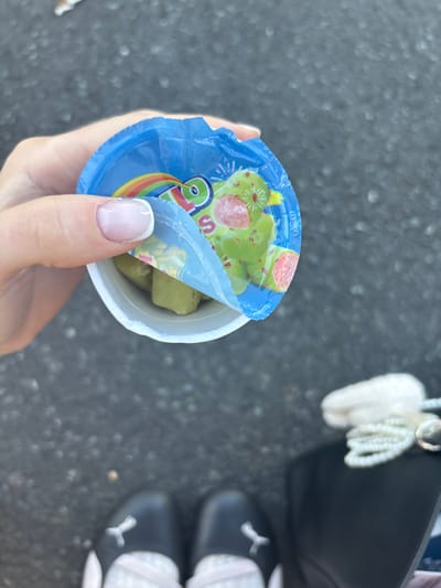 A person's hand, with a French manicure, is holding an open cup containing green food items, possibly pickles or some sort of candy. The cup has a blue lid partially peeled back, revealing a colorful design with a rainbow and some festive elements. The background appears to be an outdoor setting with a dark, asphalt-like surface. In the upper right portion of the image, a black handbag with a white fluffy charm and beaded tassel is visible, as well as a pair of dark shoes with white accents, possibly Puma brand. The overall impression is a casual, everyday moment.