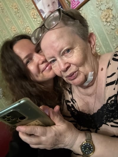 The image shows a close-up, selfie-style photograph of two women. The woman in the foreground, on the right, is an elderly Caucasian woman with short gray hair and glasses perched on her head. She is looking at the camera with a slight smile, and there is a piece of white medical tape on her neck. She is wearing a light-colored top with a black animal print and black lace trim. A thin gold necklace is visible around her neck. On her left wrist, she wears a gold-colored wristwatch with a black face and gold hands.
To her left, and slightly behind her, is a younger Caucasian woman with long, dark brown hair. She is also looking at the camera, smiling, and her cheek is touching the elderly woman's cheek. She is holding a smartphone in her right hand, which is partially visible at the bottom left of the frame. The phone has a green case and a prominent camera array on the back.
The background consists of a wall covered in green wallpaper with a floral pattern of yellow and cream-colored flowers. A framed photograph, which appears to be of a baby, is visible on the wall in the upper right portion of the image. The lighting suggests an indoor setting, possibly a home. The image does not provide any specific indicators of the city of Turinsk, Russia, or any other geographical location.