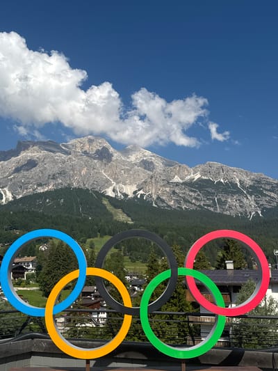 The image shows a close-up of the Olympic rings, rendered in bright, distinct colors, positioned in the foreground. The rings are made of a smooth, solid material and are interlocked in the classic arrangement. Behind the rings, a mountainous landscape unfolds under a clear blue sky dotted with fluffy white clouds. The mountains are rugged and rocky, with patches of snow visible on their peaks. A dense forest of evergreen trees covers the lower slopes, interspersed with buildings that appear to be part of a village or resort. The architecture of these buildings suggests a European setting, with dark roofs and wooden accents. The lighting indicates it is daytime, with sunlight casting shadows and highlighting the textures of the mountains and trees. The overall scene evokes a sense of natural beauty and sporting heritage, possibly in a mountainous region like the Italian Dolomites, as suggested by the architecture and terrain which are characteristic of Cortina d'Ampezzo, Italy, a known winter sports hub. There are no people or animals visible in the image.