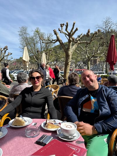 The image captures a scene at an outdoor cafe in Cochem, Germany. Two individuals are the main subjects, seated at a table covered with a pink tablecloth. A man wearing a dark blue jacket with colorful shirt underneath, sits with a light skinned male in a grey cap, while a woman in a black jacket and sunglasses sits across from them. On the table, there are plates of cake, cups with coffee, an ashtray and a red Eiskuvy Desserts menu. Behind them, there are other patrons at surrounding tables, as well as trimmed trees with bare branches, typical of a European town square. In the background, the town of Cochem and a tree-covered hillside can be seen under a clear blue sky.