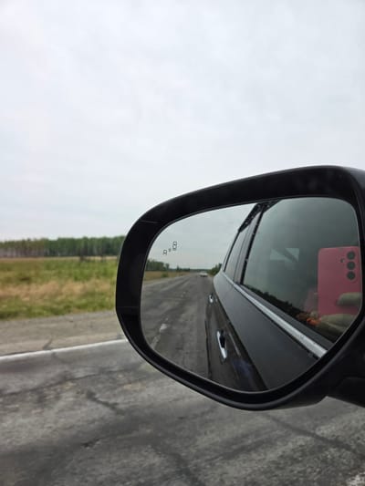 The image depicts a car's side mirror reflecting a road and surrounding landscape. A glimpse of the photographer's pink phone is visible in the mirror's reflection. The road appears to be asphalt, flanked by grassy terrain and some trees. Due to the lack of discernible landmarks, it's impossible to determine the exact location in Аур, Russia.