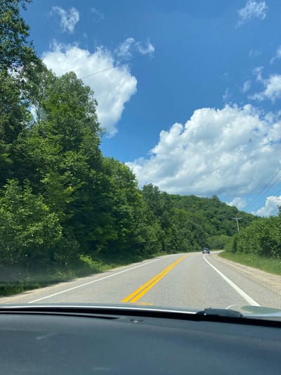 The image depicts a point-of-view shot from a vehicle traveling on a paved road that curves to the right, surrounded by lush green trees and rolling hills under a partly cloudy blue sky. The road has a white line marking the right edge and double yellow lines in the center. A dark-colored SUV is visible in the distance, traveling in the same direction. Power lines run diagonally across the sky. The foreground shows the dashboard of the vehicle from which the photo is taken. There is no indication of the specific location within the city of Lac-Simon, Canada.