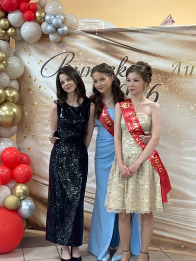 Three young women are posing for a photo in front of a decorative banner. The banner has a champagne-colored background with a swirling dark pattern and gold confetti. The words "Выпуск" (Graduation) are prominently displayed in large, elegant script.
The woman in the back, on the left, is wearing a gold, strapless, lace dress and a red sash that reads "2026" on one end and some Russian text on the other. She has her hair pulled up and is looking towards the camera with a slight smile.
The woman in the middle is wearing a light blue dress and a similar red sash with Russian text. Her dark hair is down, and she is also looking towards the camera with a neutral expression.
The woman in the front, on the right, is wearing a long, black, sequined dress. Her dark hair is down, and she is looking directly at the camera with a bright, engaging smile. She is holding a smartphone in her right hand.
To the left of the women, a balloon arch is visible. It is composed of white, silver, red, and gold balloons of various sizes. To the right of the women, more balloons are visible, including large red and white ones, along with gold and silver accents.
The lighting suggests it is daytime, and the indoor setting is likely a venue decorated for a graduation event. The overall mood appears celebratory and joyful.