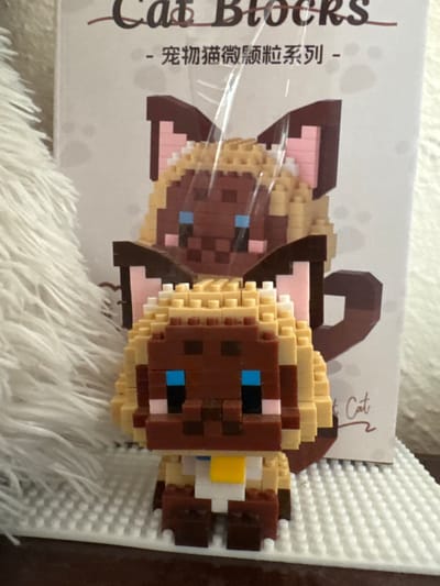 The image shows a close-up of two Siamese cat figures made of small building blocks, possibly from a Nanoblock or similar brand. The figures are positioned in front of a cardboard box that has Chinese and English text, including "Cat Blocks" and what appears to be a brand name. The box also features paw prints and an outline of a cat's head. The foreground figure is a Siamese cat with brown fur, tan ears, blue eyes, and a yellow collar. The background figure, partially obscured by the foreground figure and a clear plastic wrapper, is also a Siamese cat with similar coloring. The figures are placed on a white base plate made of the same building block material. To the left of the figures, a fluffy white material, possibly a rug or blanket, is visible. The lighting suggests an indoor setting. The image is focused on the toys and their packaging. There is no information in the image to indicate a location in the city of Mamaivtsi, Ukraine.