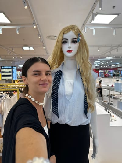 A young woman with dark hair pulled back and wearing a black garment and a white seashell necklace is posing for a selfie next to a female mannequin in a store. The mannequin has blonde wavy hair and is wearing a light blue blouse and a black skirt. The background appears to be a retail store with clothing racks and displays. The woman is smiling at the camera. The scene is well-lit, likely due to overhead store lighting.