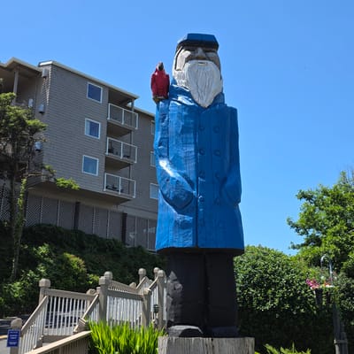 This image captures a towering wooden sculpture of a fisherman in Cannon Beach, Oregon, USA. The sculpture, painted in vibrant blue and black, depicts a man with a white beard, wearing a cap and a coat with visible buttons. A small red bird is perched on his shoulder. In the background, a multi-story building with gray siding and balconies is visible, nestled amongst lush green trees and foliage. A white wooden railing and stairs lead up towards the building, with some greenery in the foreground. The sky above is a clear, bright blue.