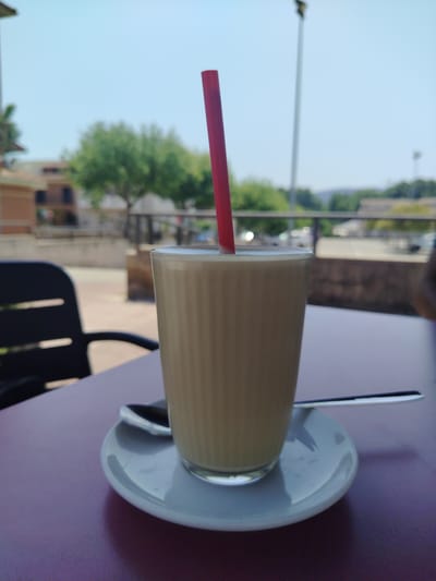 A tall, ribbed glass filled with a light-colored beverage, possibly coffee or a milkshake, sits on a white saucer on a dark purple table. A red straw extends from the drink. A silver spoon rests on the saucer. In the background, out of focus, is a black chair. Beyond the chair, a tree-lined street with buildings and parked cars is visible under a clear blue sky. The location is identified as Pla Del Temple, Spain.