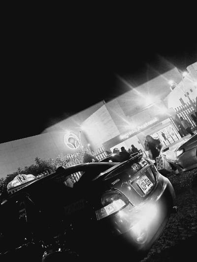 The black and white image depicts a nocturnal urban scene in front of the MegaPlaza Chimbote shopping mall in Chimbote, Peru. The foreground is dominated by the rear of a dark-colored sedan with a spoiler, displaying Peruvian license plate V2P-036. To the left, a portion of another vehicle, identifiable as a taxi by a roof-mounted "TAXI" sign with an encircled "N", is visible. In the midground, multiple blurred human figures are present, some appearing to walk, with at least one individual on the right carrying bags. Another lighter-colored vehicle is partially visible on the far right. The background features the brightly illuminated facade of the MegaPlaza Chimbote building, showcasing its prominent logo and name. A sign for "Banco de la Nación" is also discernible. Streetlights or building lights create noticeable lens flares throughout the upper portion of the scene. The overall environment appears to be an outdoor area, likely a parking lot or street, adjacent to the mall entrance.