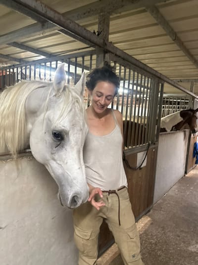 The image depicts an indoor stable in Kfar Monash, Israel. A fair-skinned female with dark hair styled in a bun, wearing a light-colored tank top and khaki cargo pants, stands in the foreground, looking down and to her left with a smile. Her left hand is raised near the muzzle of a light-grey or white horse. This horse's head and neck are visible, extending out of an open stall. The stall features a solid light-colored lower wall and vertical metal bars above. In the background, another horse with a brown and white pinto coat is partially visible within a separate stall, behind metal bars. The stable ceiling consists of light-colored corrugated metal sheets and support beams. The floor is an uneven concrete or packed earth surface. A dark rope hangs on a partition to the right of the female.