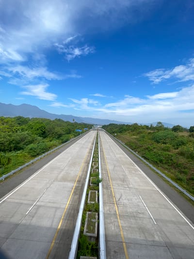 An aerial, wide-angle shot captures a multi-lane highway stretching into the distance, flanked by lush green vegetation and distant mountains under a bright blue sky with scattered clouds. The highway, composed of concrete sections, is divided by a median strip with planters containing greenery. Yellow and white lines delineate the lanes. In the background, a tall tower is visible amidst the trees, and further in the distance, rolling mountains form the horizon. The location is identified as the Seulimeum Toll Road in Seulimeum, Indonesia. There are no people or vehicles visible in the image.